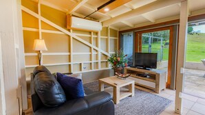 A living room with a sofa, a coffee table, a TV, and a window overlooking a grassy area. - The Dairy @ Cavan, Kangaroo Valley, Stunning Views (Barrengarry)