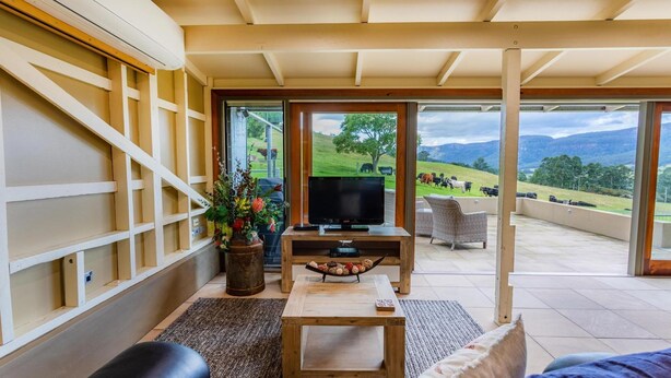 A living room with a TV, a wooden coffee table, a sofa, and a large window offering a view of a farm and hills. - The Dairy @ Cavan, Kangaroo Valley, Stunning Views (Barrengarry)