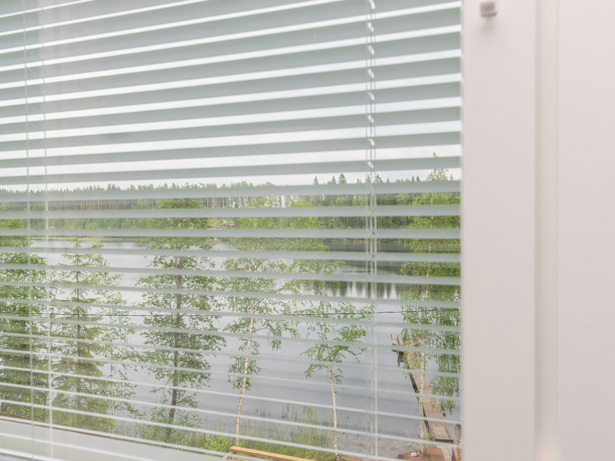 A window with horizontal blinds, through which a view of a water body and trees is visible.