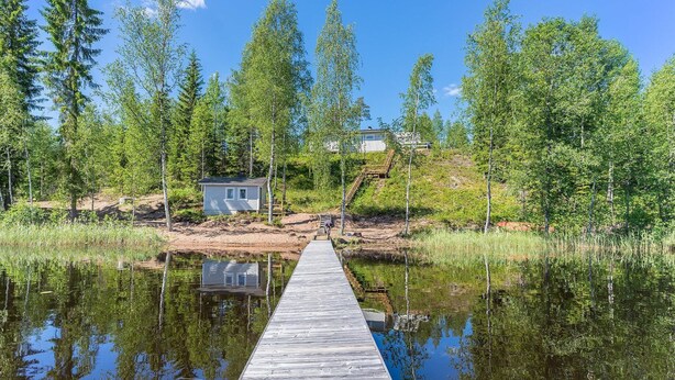 A wooden dock extending into a calm lake with reflections of trees and a small cabin in the background. - Valonsäde by Interhome (Valkeala)