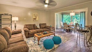 A living room with a brown sofa, a glass coffee table, and teal ottomans. There is a dining area with patterned chairs and a table. - Lake O’ the Pines Family Getaway (Avinger)