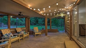 A covered patio area with wooden chairs and a table, string lights overhead, and a view of a wooded area. - Lake O’ the Pines Family Getaway (Avinger)