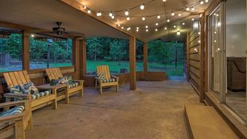 Ein überdachter Patio-Bereich mit Holzstühlen und einem Tisch, Lichterketten darüber und Blick auf ein Waldstück.