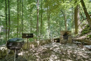 A forested area with a stone barbecue grill and metal chairs. - 'Snuggle Inn' in the Heart of Chalet Village! (Gatlinburg)