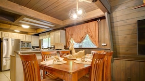 Uma mesa de jantar de madeira com seis cadeiras, um lustre e uma cozinha com armários de madeira e uma geladeira.