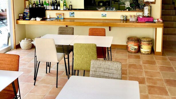 A restaurant with a ceiling fan, a thatched roof, a counter with various bottles, a kitchen in the background, and several tables and chairs. - Asobi Base Yamabare House Ishigakijima (Ishigaki)