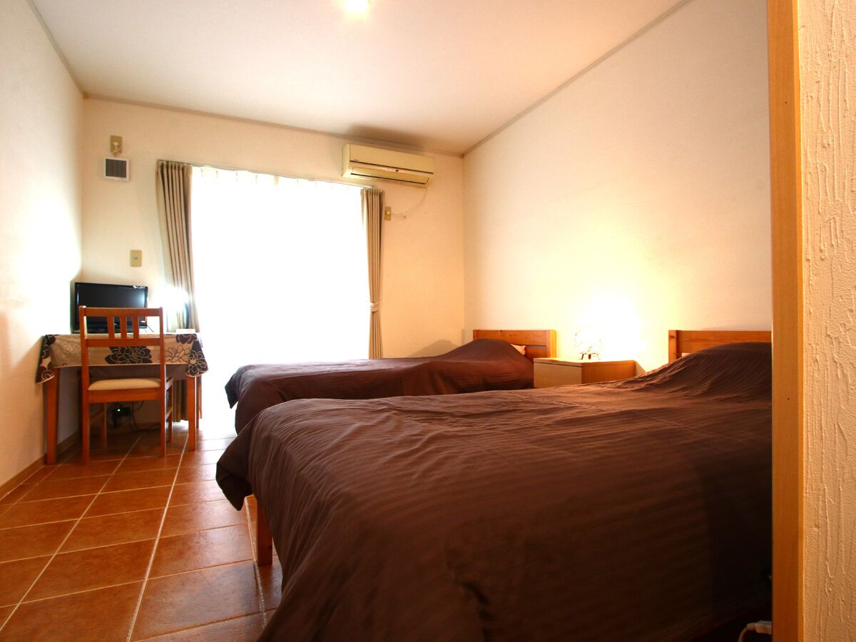 A room with two beds, a desk, a chair, an air conditioning unit, and a window with curtains.