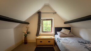 Une petite chambre mansardée, chaleureuse et accueillante, avec un plafond en pente, une fenêtre avec des rideaux, une table de chevet en bois, un lit recouvert d’une couvre-lit à motifs et un vase rempli de fleurs.