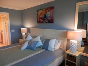 A bedroom with a bed, two bedside tables with lamps, a colorful abstract painting on the wall, and a mirror on the wall. - OV 317 Golf/Pond View Condo - Paradise Vacation Rental in Ocean Village (FORT PIERCE)