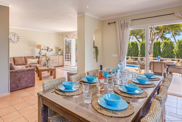 A dining area with a long table set for six, surrounded by wicker chairs, with a view of the outdoor patio.