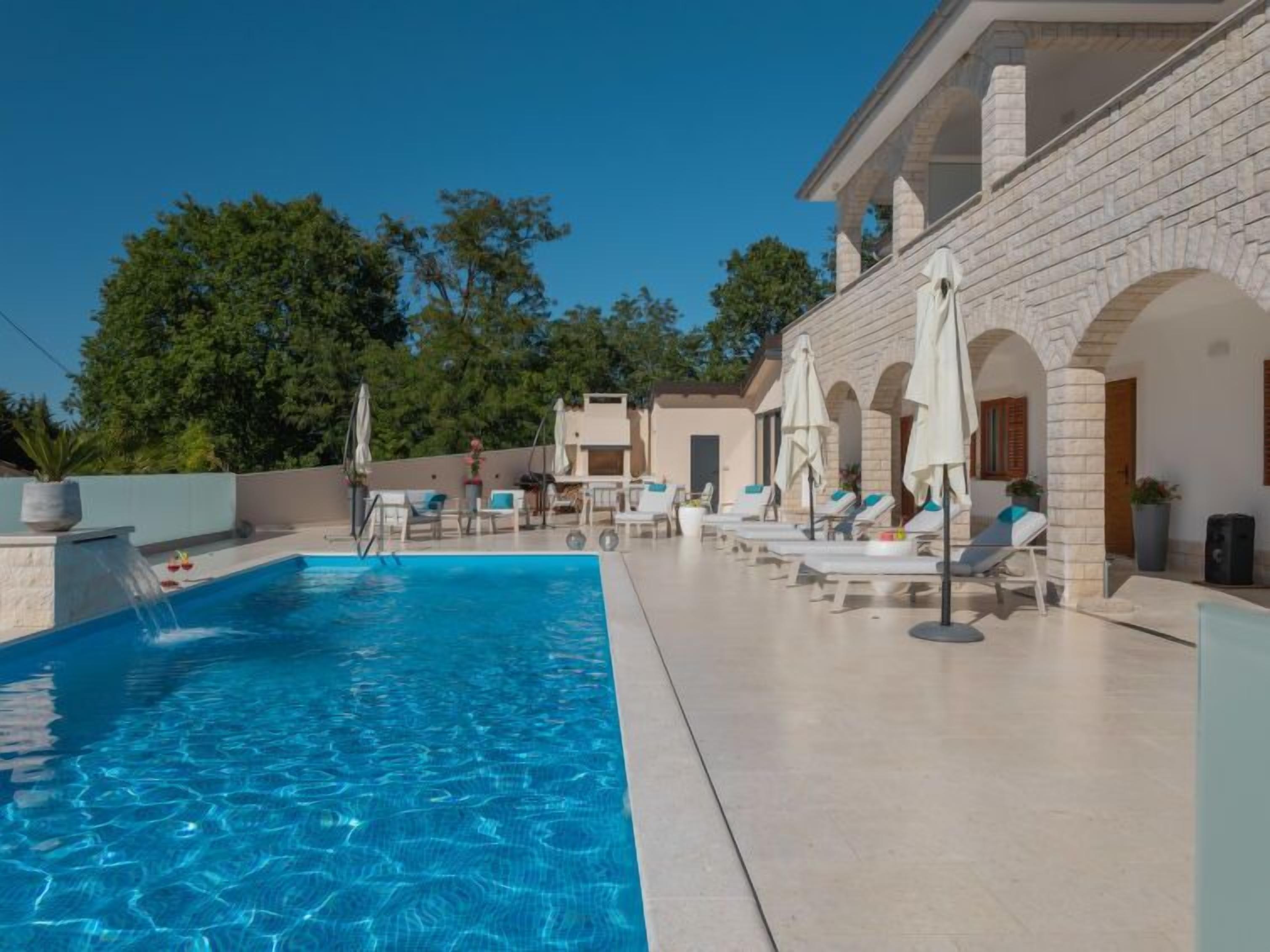 A swimming pool with lounge chairs and umbrellas, surrounded by a building with arches and a clear sky.