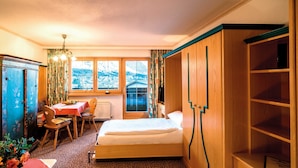 Una camera da letto con un letto, un'armadio in legno, una scrivania con una sedia, un tavolino con una ciotola di frutta e una finestra vista casa e montagne.