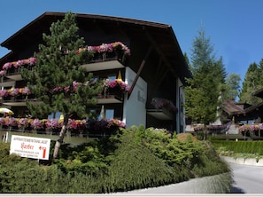 A multi-story building with balconies adorned with flowers, a sign indicating it is an apartment complex, and a clear sky in the background. - 1-room apartment (approx. 32 m²) - Apartment complex Kerber Tirol (Seefeld in Tirol)