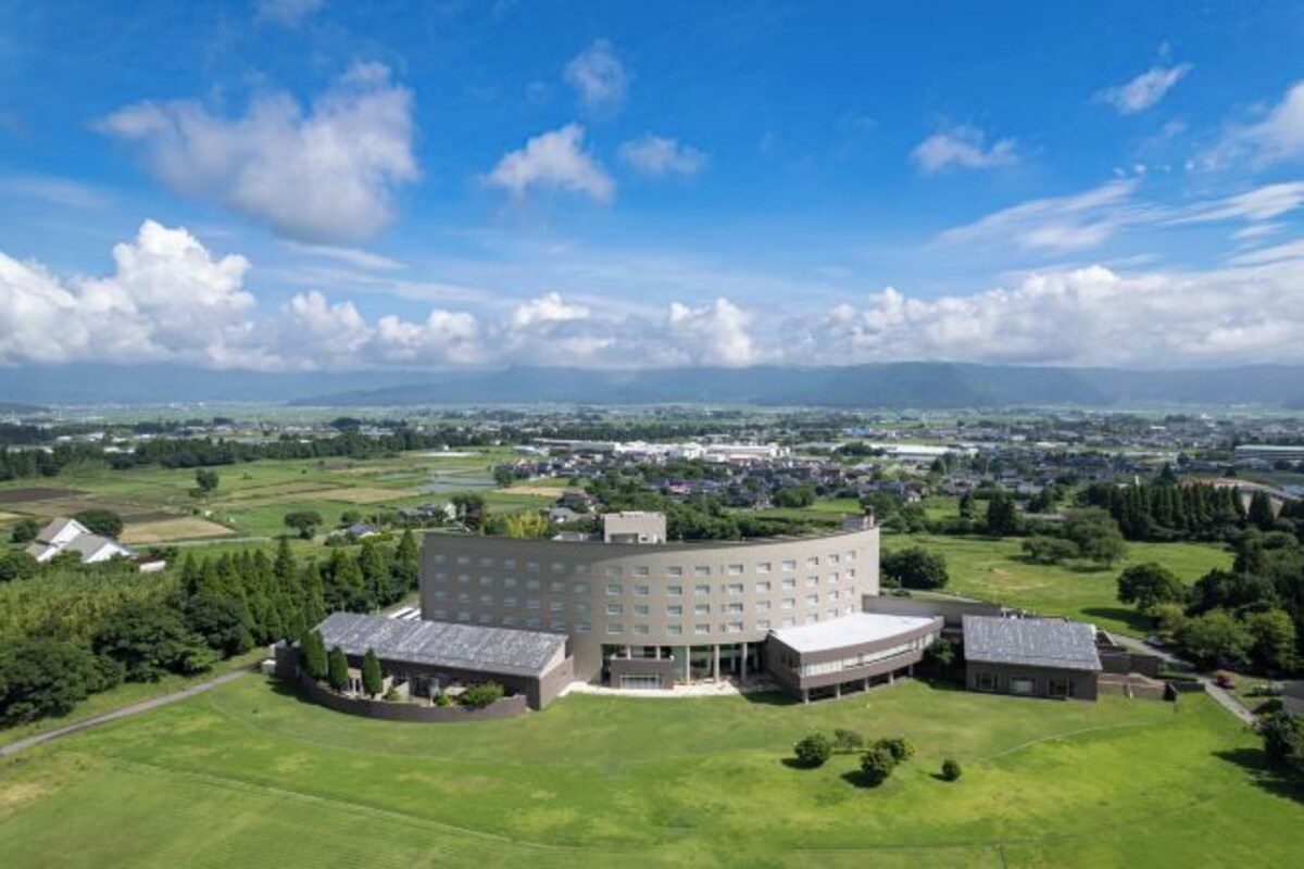 A large, modern building with a central courtyard, surrounded by green fields and a clear sky.