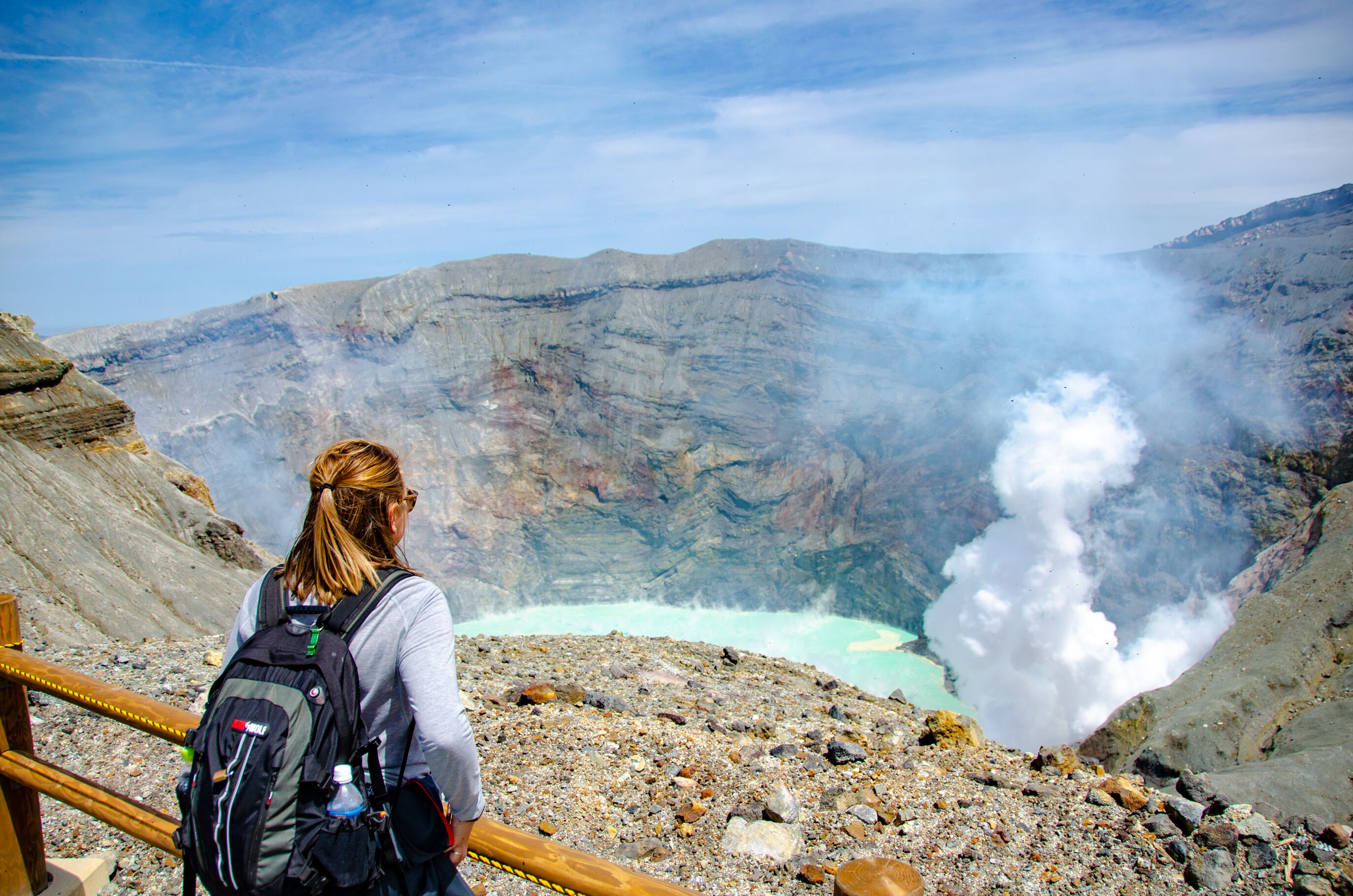A person with a backpack overlooking a mountainous landscape with a visible geothermal feature.