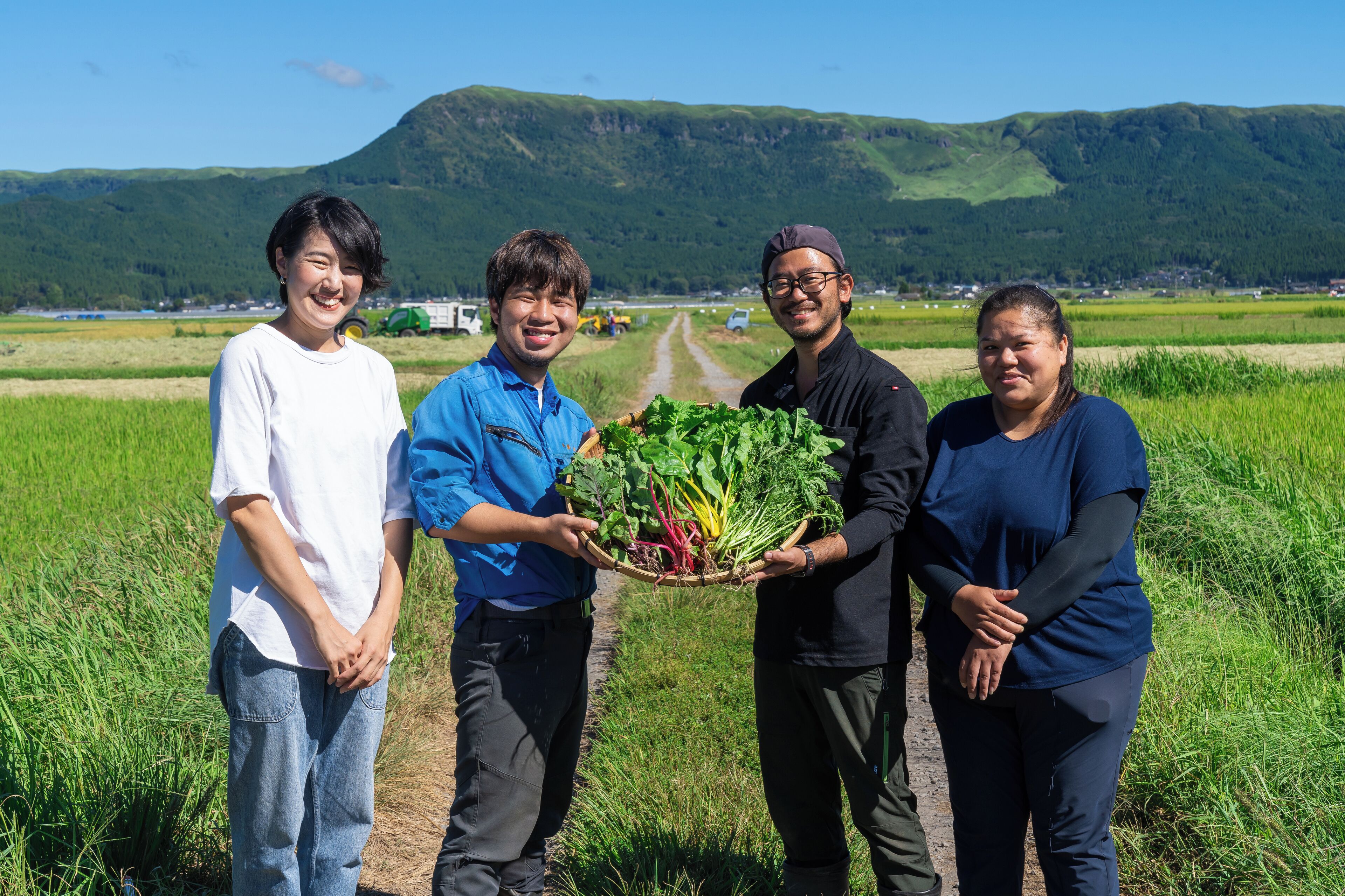 Four people holding a basket of vegetables in a rural setting with a dirt path and green fields.