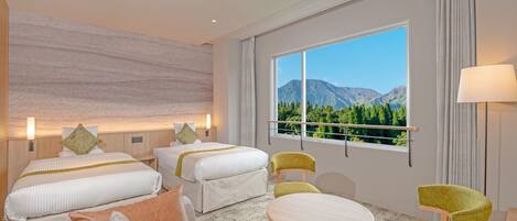 A hotel room with two beds, a sofa, a small table, and chairs. There is a large window with a view of mountains.
