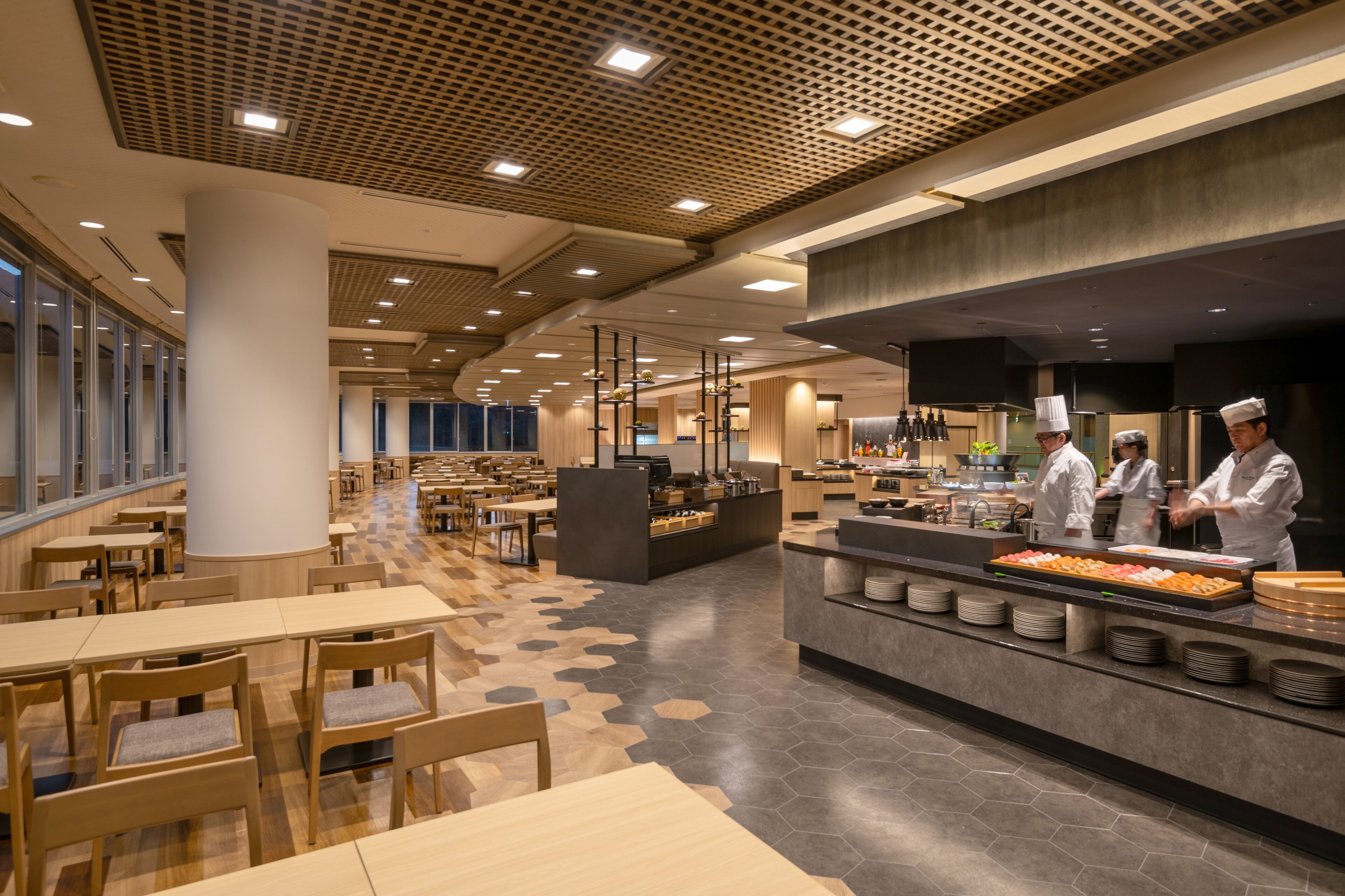 Una cafetería moderna con mesas y sillas de madera, un mostrador de buffet con comida y una zona de cocina con cocineros.