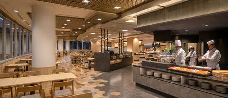 Una cafetería moderna con mesas y sillas de madera, un mostrador de buffet con comida y una zona de cocina con cocineros.