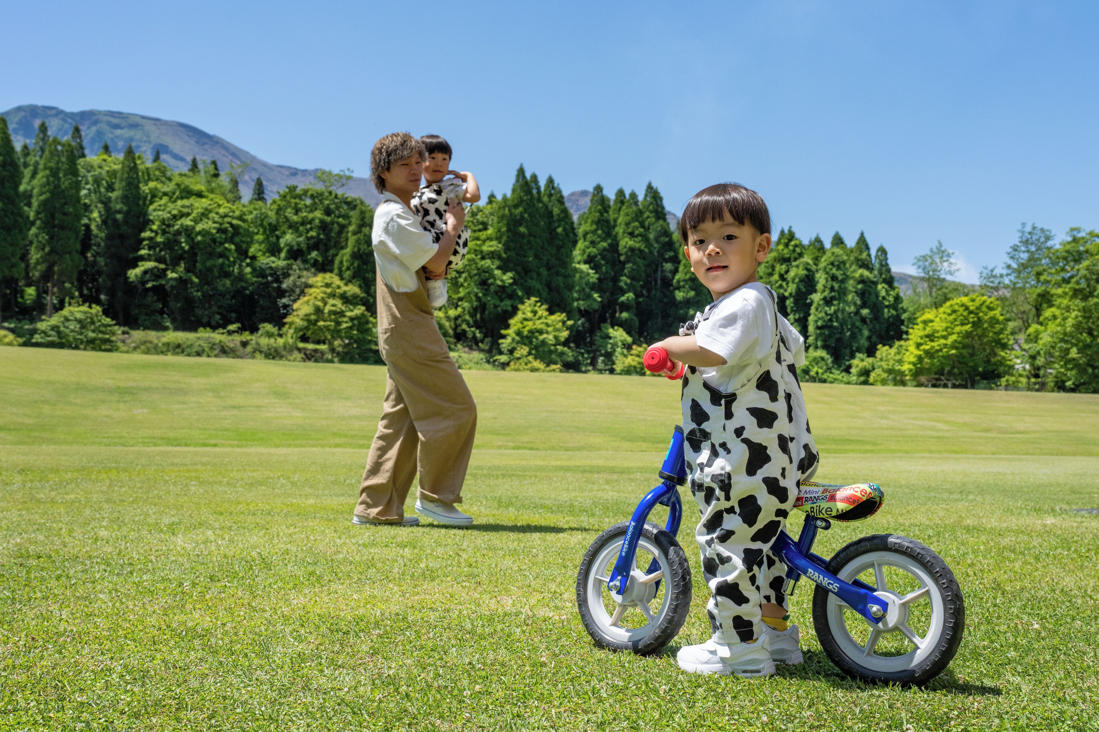 Un champ verdoyant où l’on voit un enfant sur un tricycle bleu et un adulte qui tient un enfant dans ses bras.