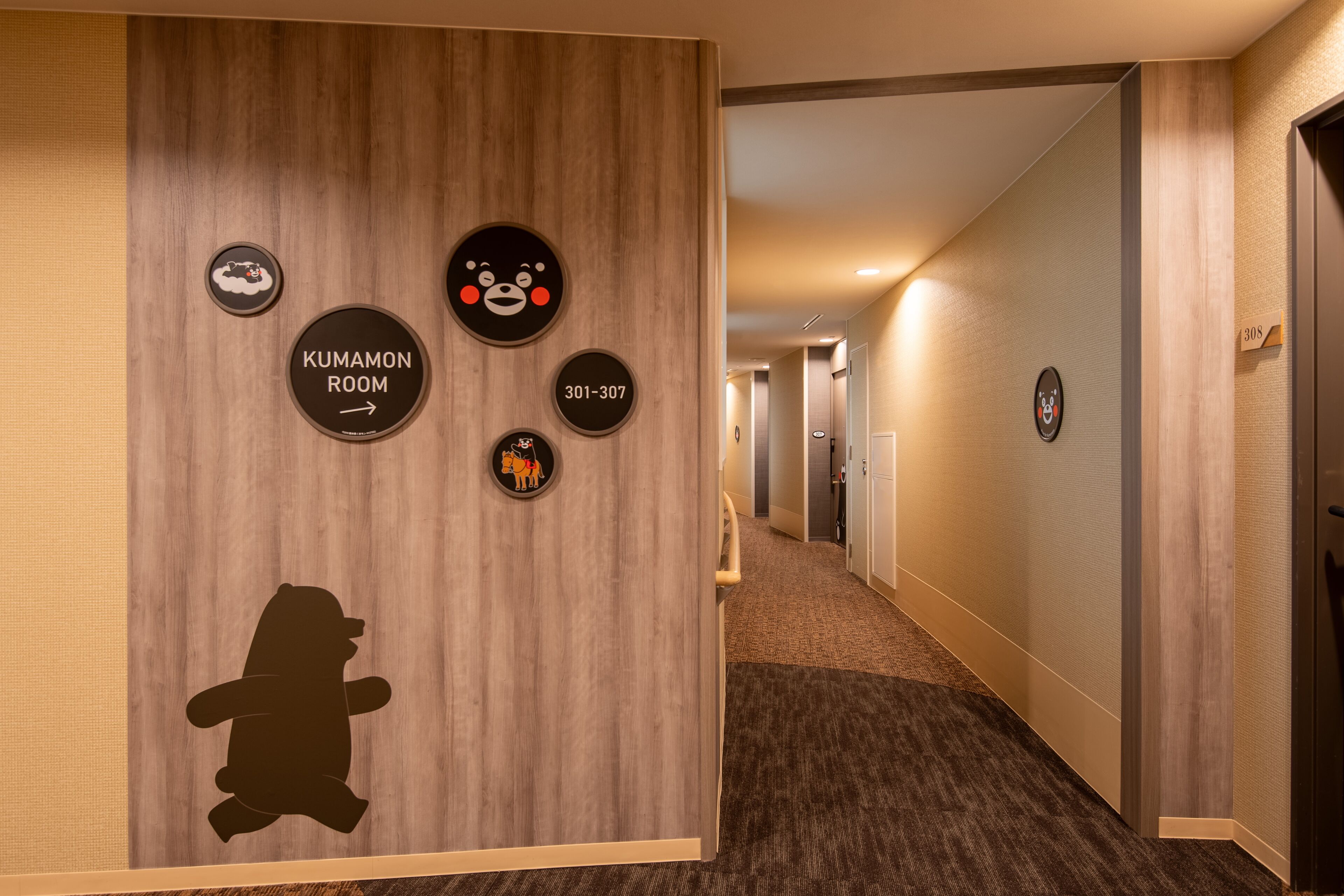 A hallway with a wooden wall featuring circular signs for 'KUMAMON ROOM' and a phone number, a silhouette of a bear, and a door labeled '30S'.