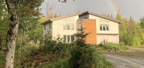 A modern two-story house with a large front porch, surrounded by dense greenery and a visible rainbow in the background. - 4 Master Suites in the Heart of Alaska...Abby's Place..... (Palmer)