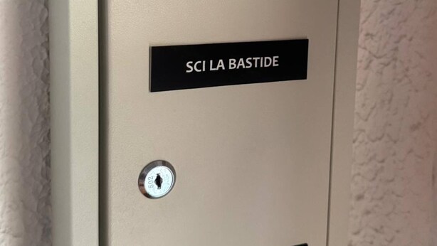Un boîtier d’interphone beige, sur lequel est apposée une plaque indiquant « SCI LA BASTIDE », et une clé y est accrochée.