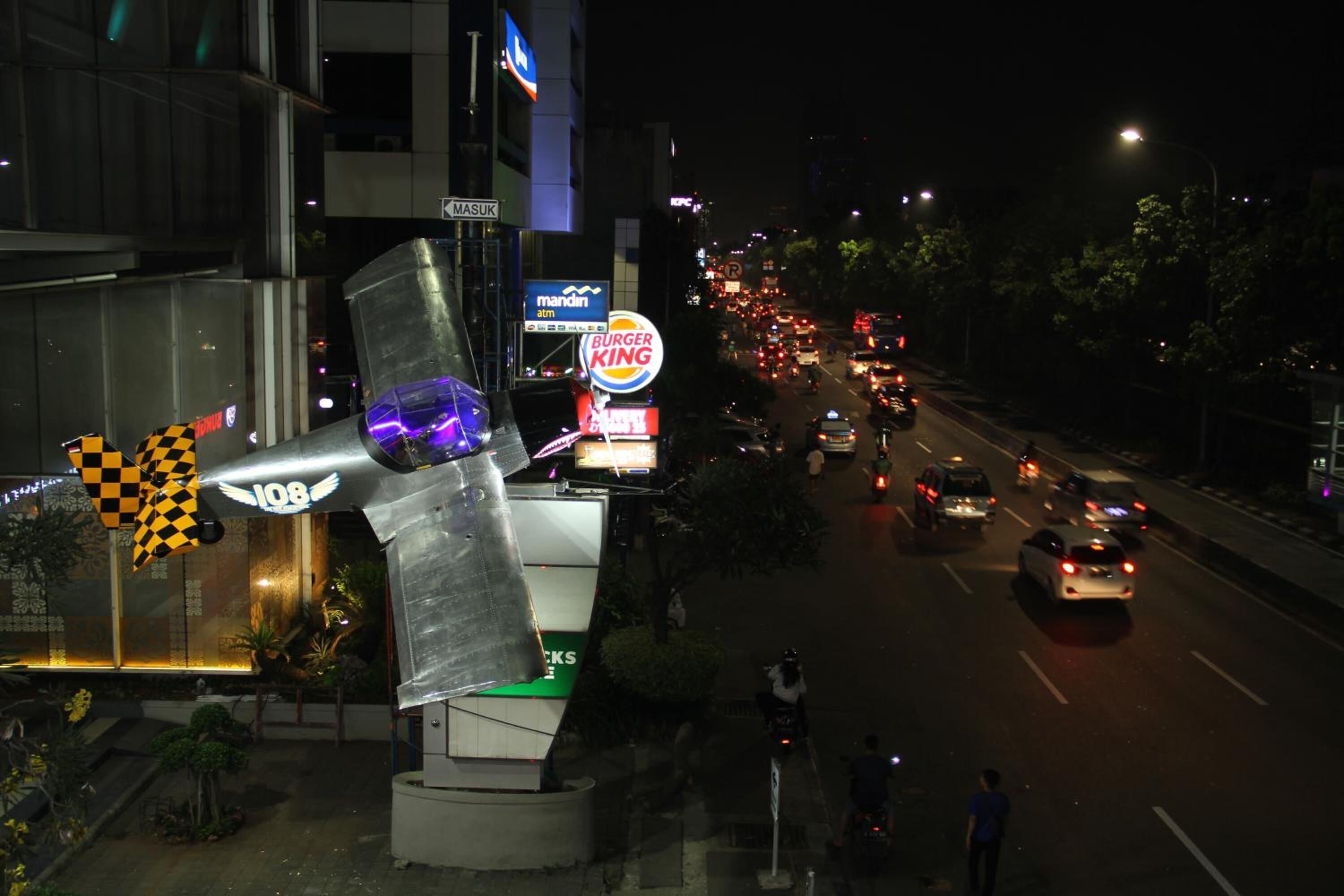 A street view at night with a Burger King sign, a large sculpture, and a MASUK sign.