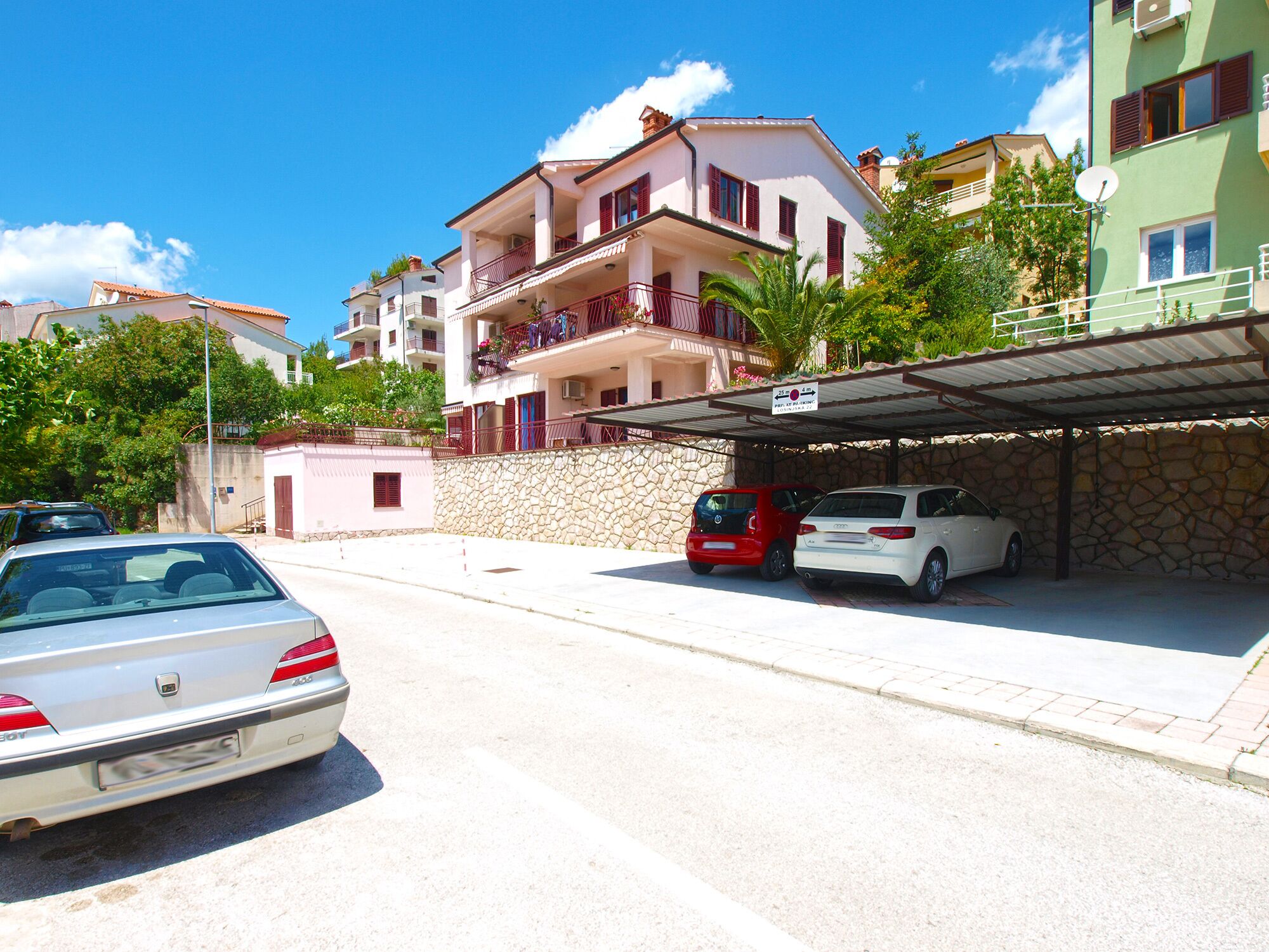 A street view with a white car parked on the side, a red car in a covered parking area, and a building with balconies and a stone wall.