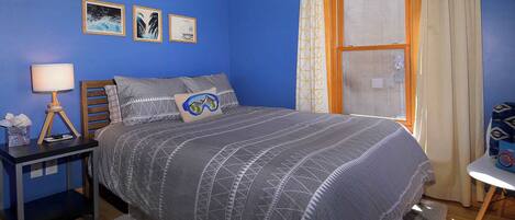 Ein Schlafzimmer mit Bett, Nachttisch, Lampe, Fenster mit Vorhängen und Wandbildern.