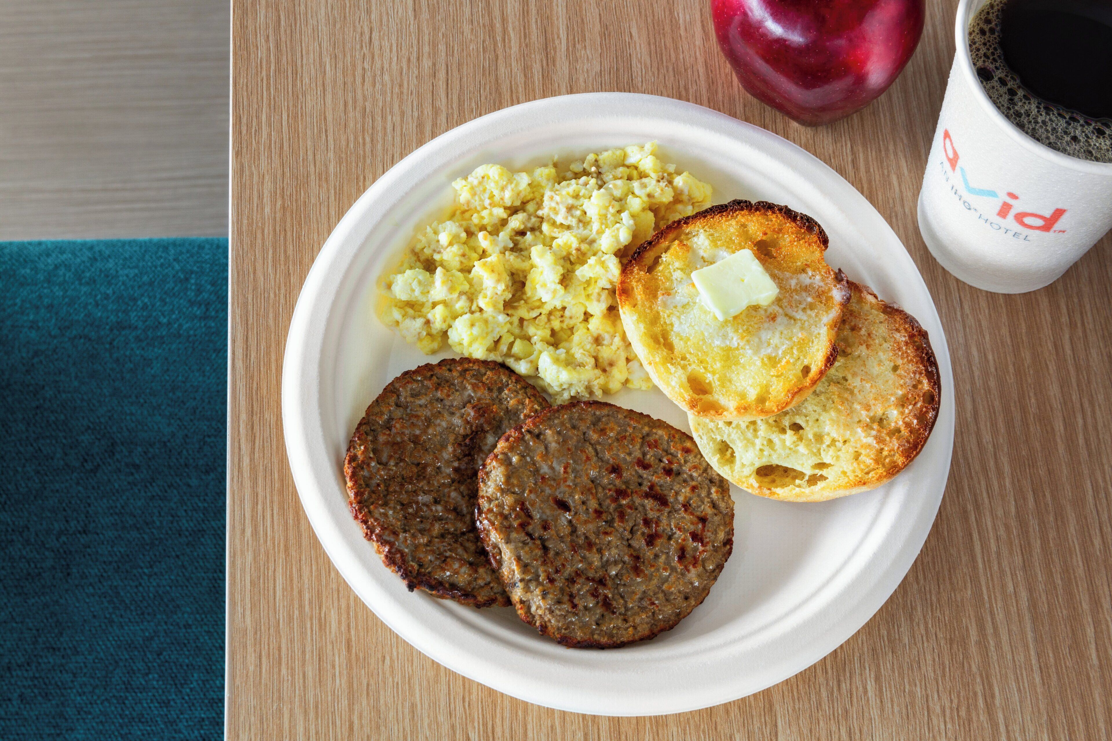 A plate with scrambled eggs, toast, and two patties, a red apple, and a coffee cup on a wooden table.