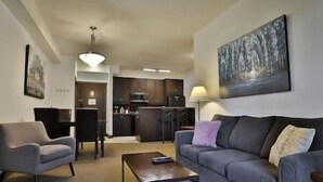 Ein Wohnzimmer mit grauem Sofa, einem Couchtisch und einem Sessel. Im Hintergrund ist eine Küche mit Schränken und einem Kühlschrank zu sehen.