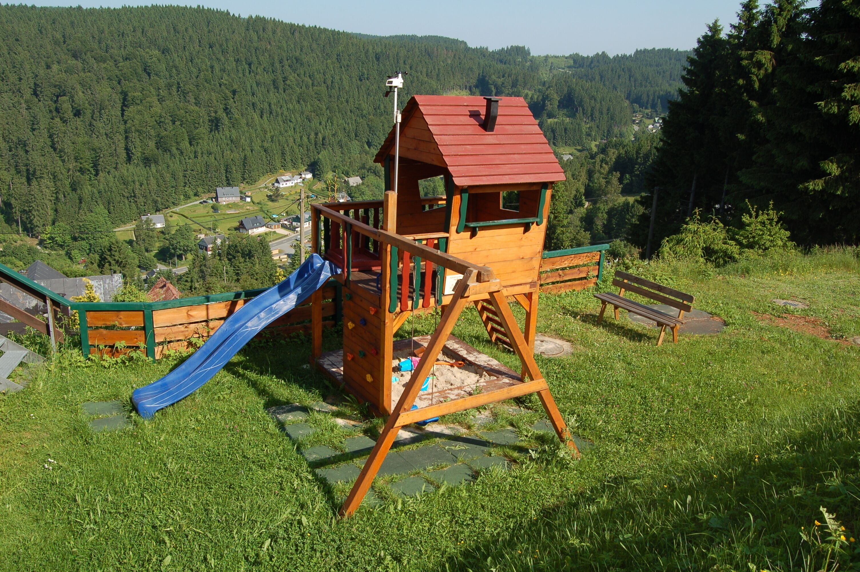 A wooden play structure with a red roof and a blue slide, surrounded by green grass and a forested hillside.