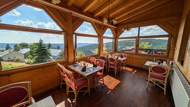 A wooden dining area with red cushioned chairs and tables, large windows offering a view of a forested landscape.