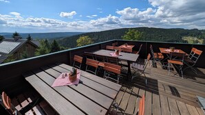 A wooden deck with tables and chairs, overlooking a forested valley.