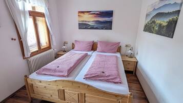 A double bed with checkered bedding, a bedside table with a lamp, a window with curtains, a radiator, and two landscape paintings on the walls.