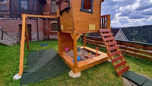 A wooden playhouse with a slide and a small sandbox, set against a brick building with a view of a forested hill.