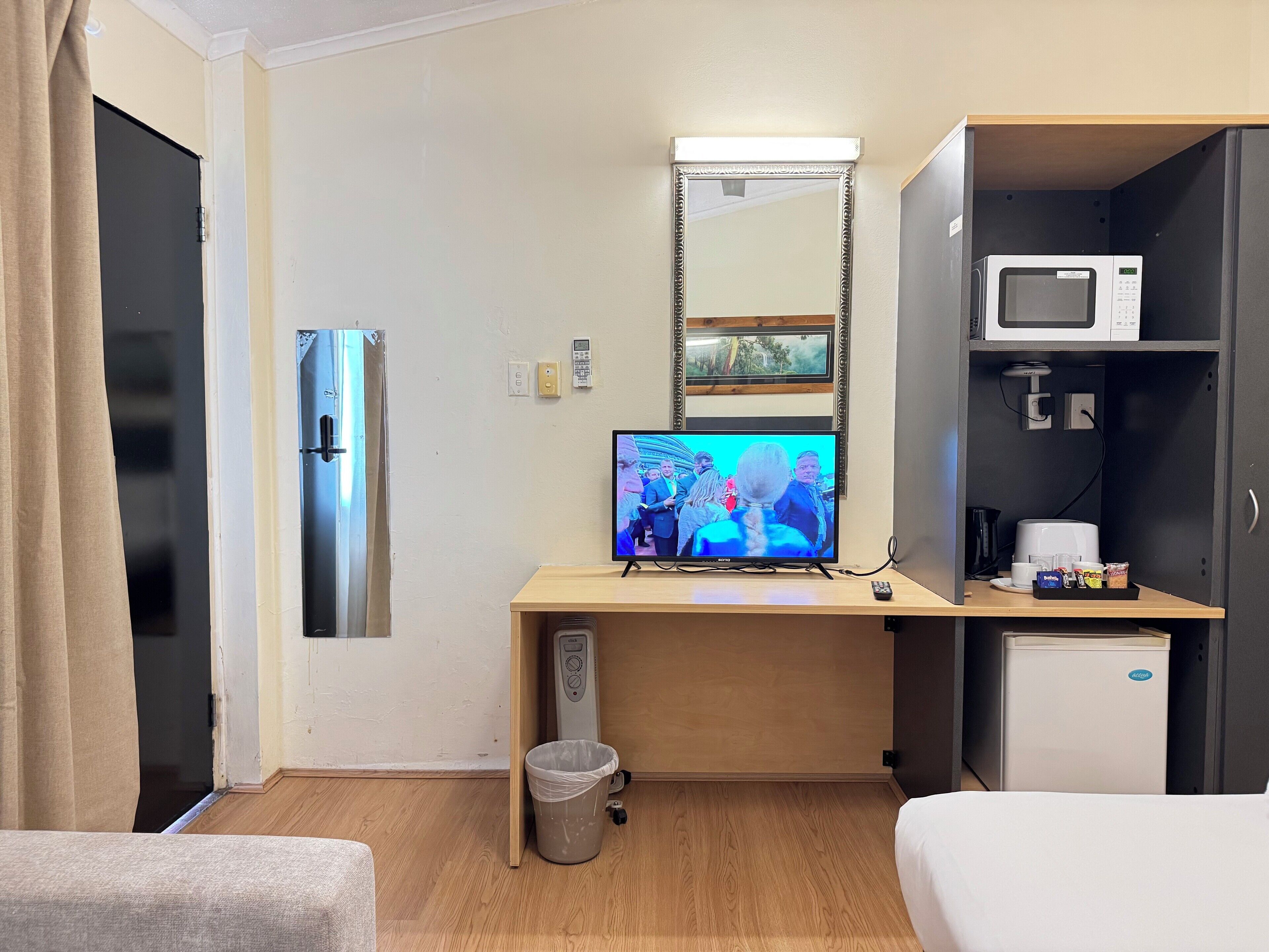 Una camera d'albergo con un letto, una scrivania con televisore, un frigorifero, un forno a microonde, un piccolo frigo, un cestino e un comodino con telecomando.