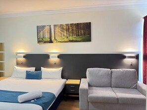 A hotel room with a large bed, a grey armchair, a nightstand, and a painting of a forest on the wall. - Taree Motor Inn (Taree)