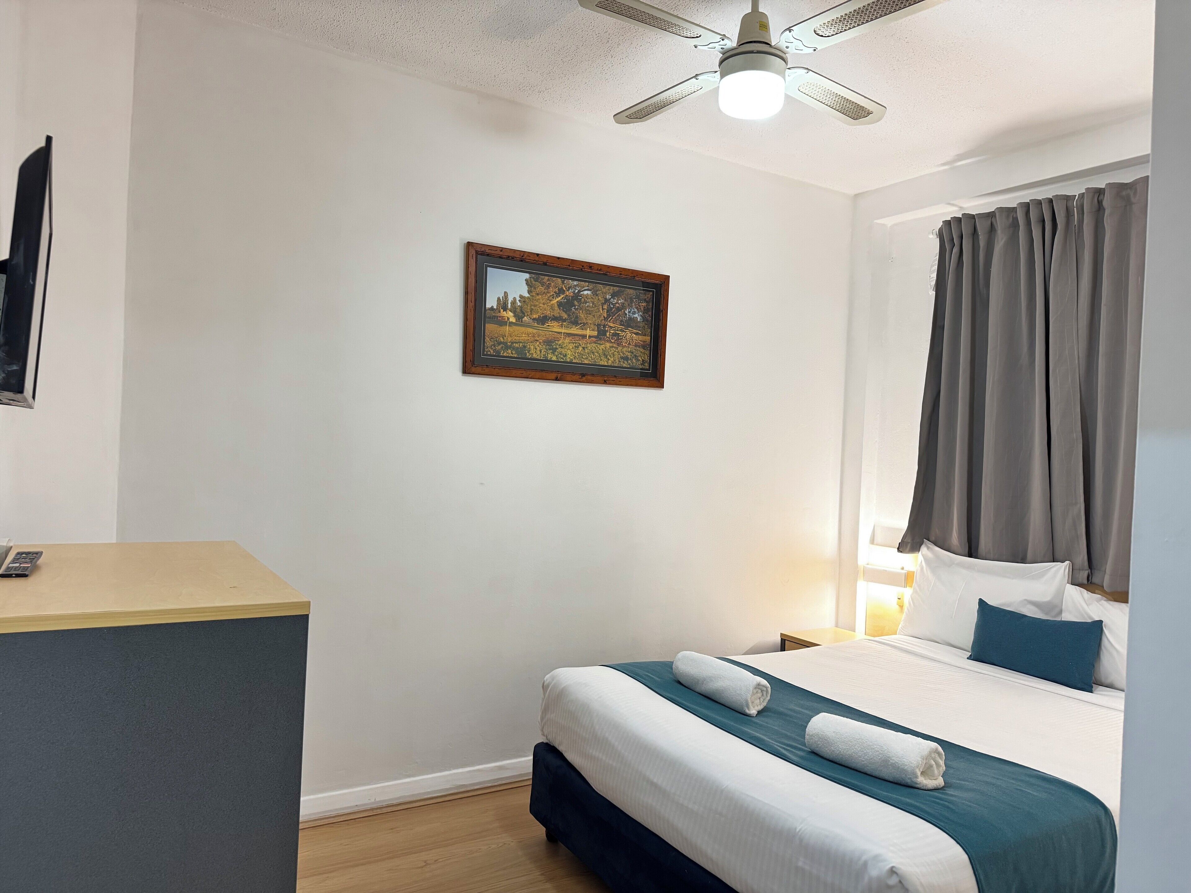 Una camera d'albergo con un letto, una scrivania, una ventola a soffitto, un quadro appeso al muro e una finestra con tende.