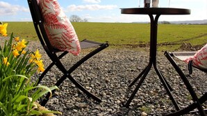 A table with two chairs and a bottle of wine, set on a gravel surface with a grassy field in the background. - The Shearer's Hut, Sleeps 2, Wood Burner, Fire Pit (Carmarthen)