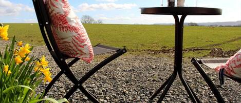 A table with two chairs and a bottle of wine, set on a gravel surface with a grassy field in the background.