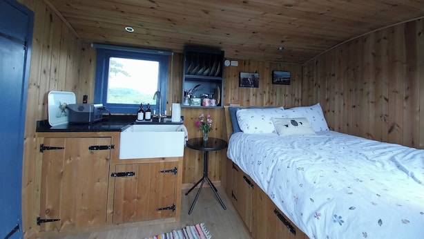 A wooden cabin interior with a bed, a small table, a sink, and a window with a view. - The Shearer's Hut, Sleeps 2, Wood Burner, Fire Pit (Carmarthen)