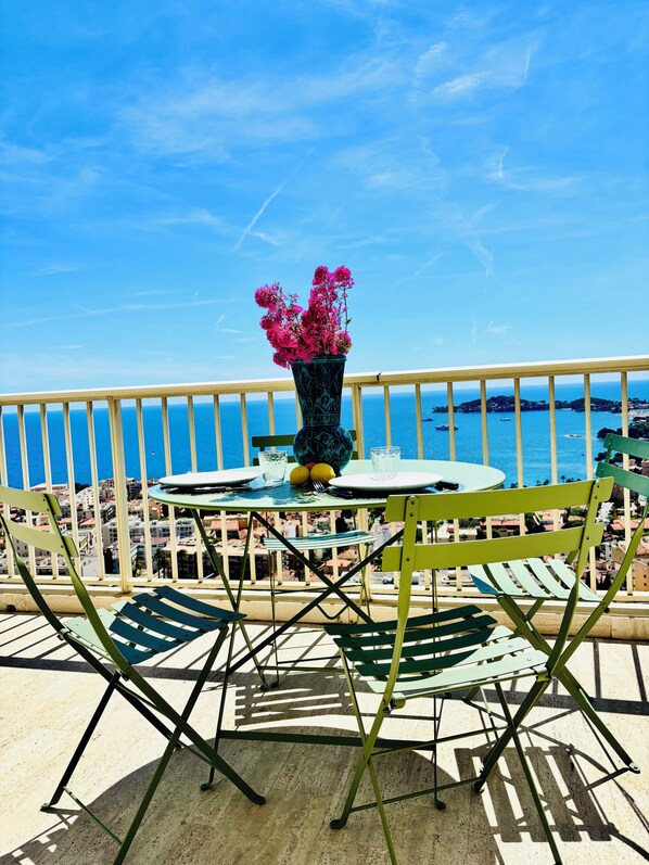 A balcony with a table, chairs, a vase with flowers, and a view of the sea.