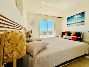 A bedroom with a bed, a wicker chair, a sofa, a view of the city, and a balcony.