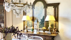 A dining area with a table, chairs, a chandelier, lamps, a painting, and a mirror.