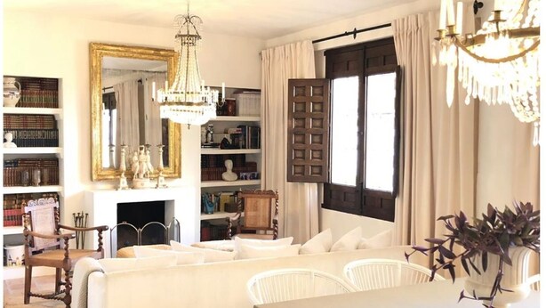 A cozy living room with a fireplace, a large mirror, and a chandelier. There are bookshelves filled with books and decorative items.