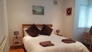 A neatly made bed with a white duvet, two dark pillows, and two folded towels on the bed. There is a small bedside table with a lamp on each side of the bed. The room has wooden flooring, a radiator, and a window with blinds. - Atlantic Retreat - 5 minute stroll from Crooklets Beach (Bude)
