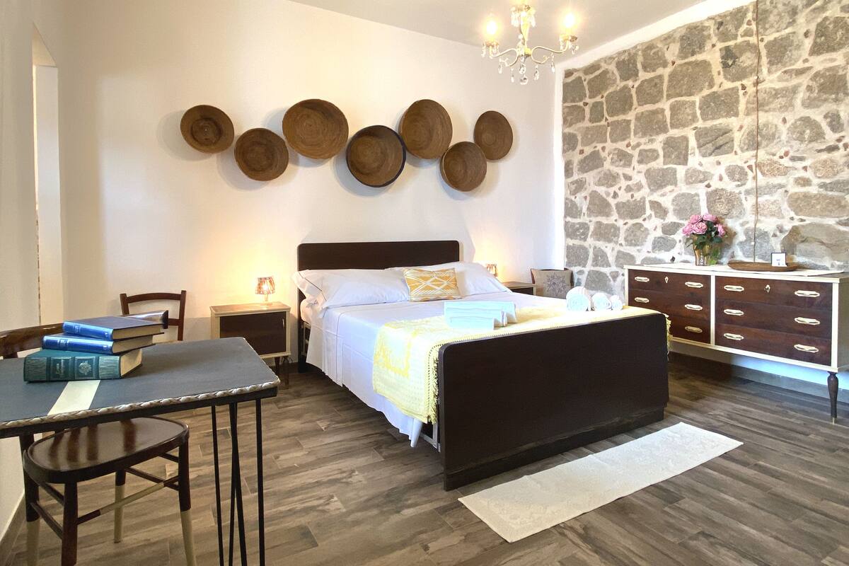 A bedroom with a bed, bedside tables, a dresser, a chandelier, and a stone wall.