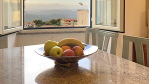 A dining table with a fruit bowl containing apples, bananas, and oranges, positioned near a window with a view of a cityscape and mountains. - Canduleri (Orgosolo)